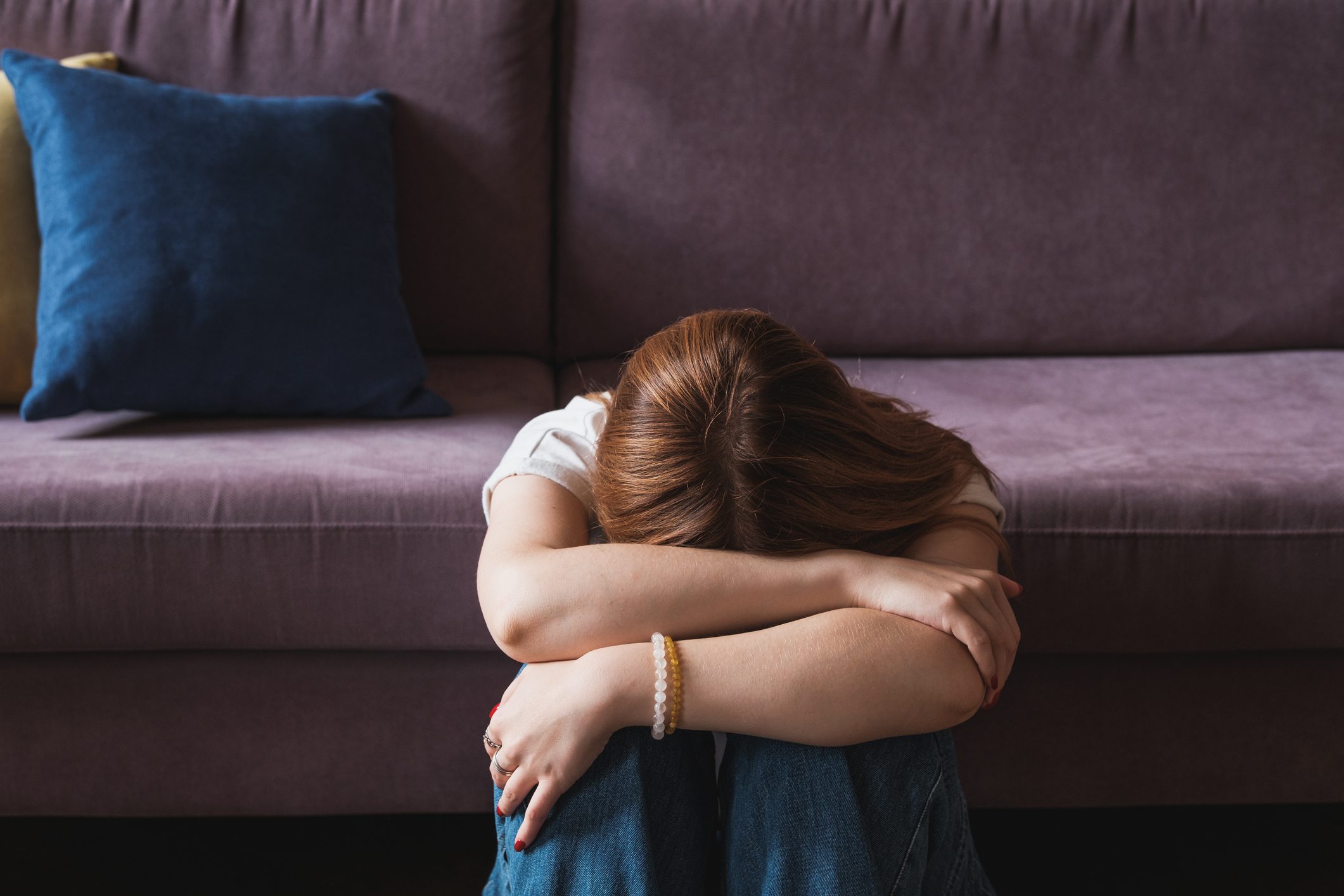 Upset depressed woman covers her face with her hands and sits on the floor at the edge of her sofa.